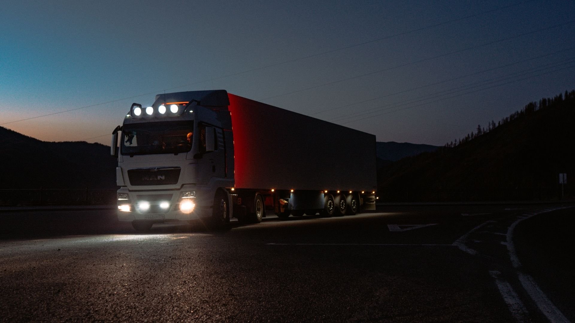 Large semi-truck with red cargo container driving on mountain road at twilight with headlights on.