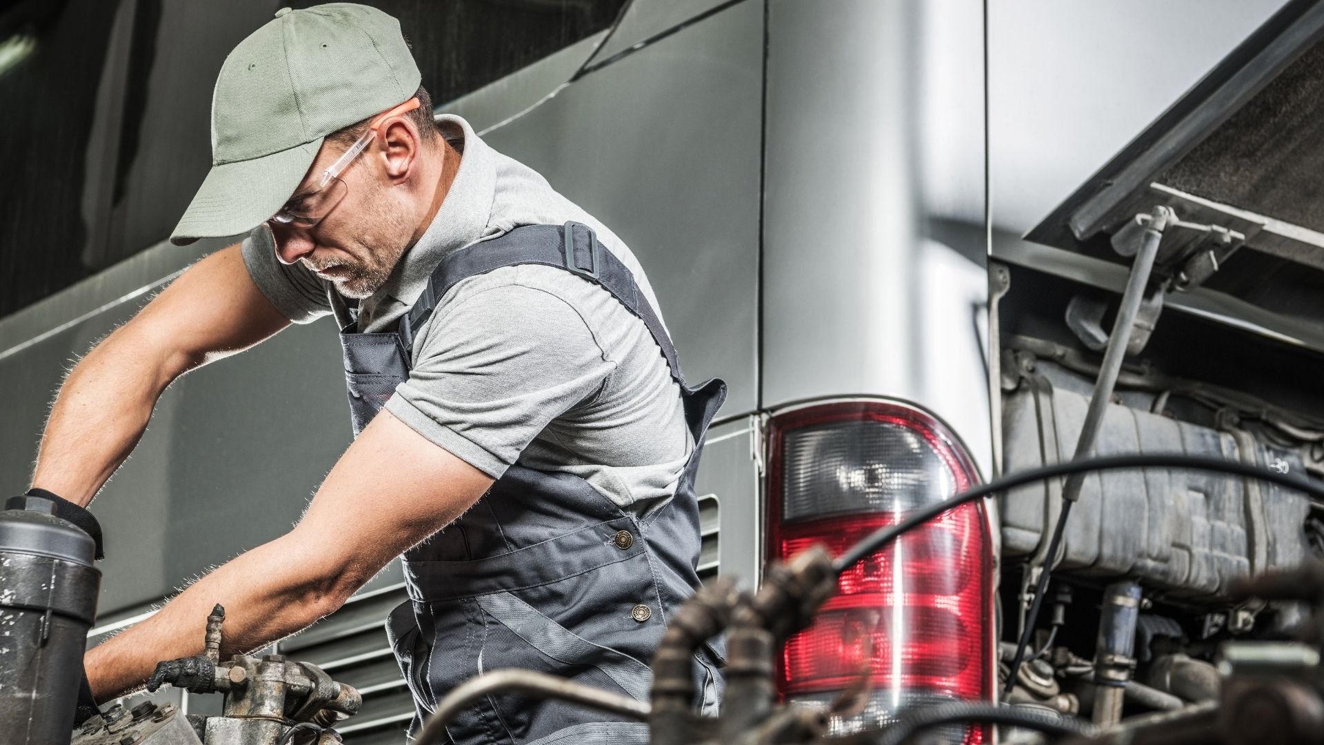 Mechanic working on vehicle engine with tools and protective gear near taillight.
